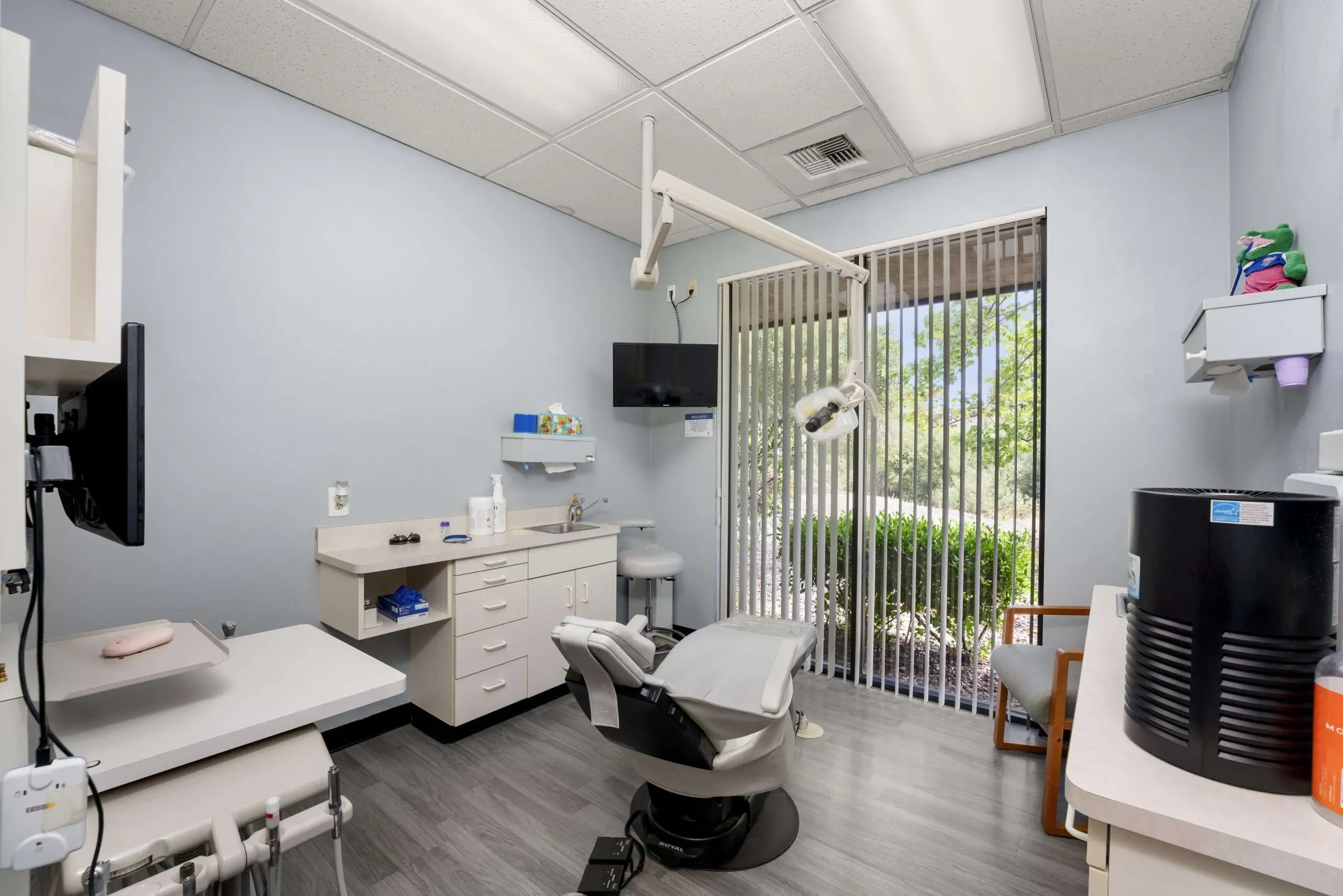 Treatment room with a dental chair, overhead light, monitor and equipment cabinets beside a window