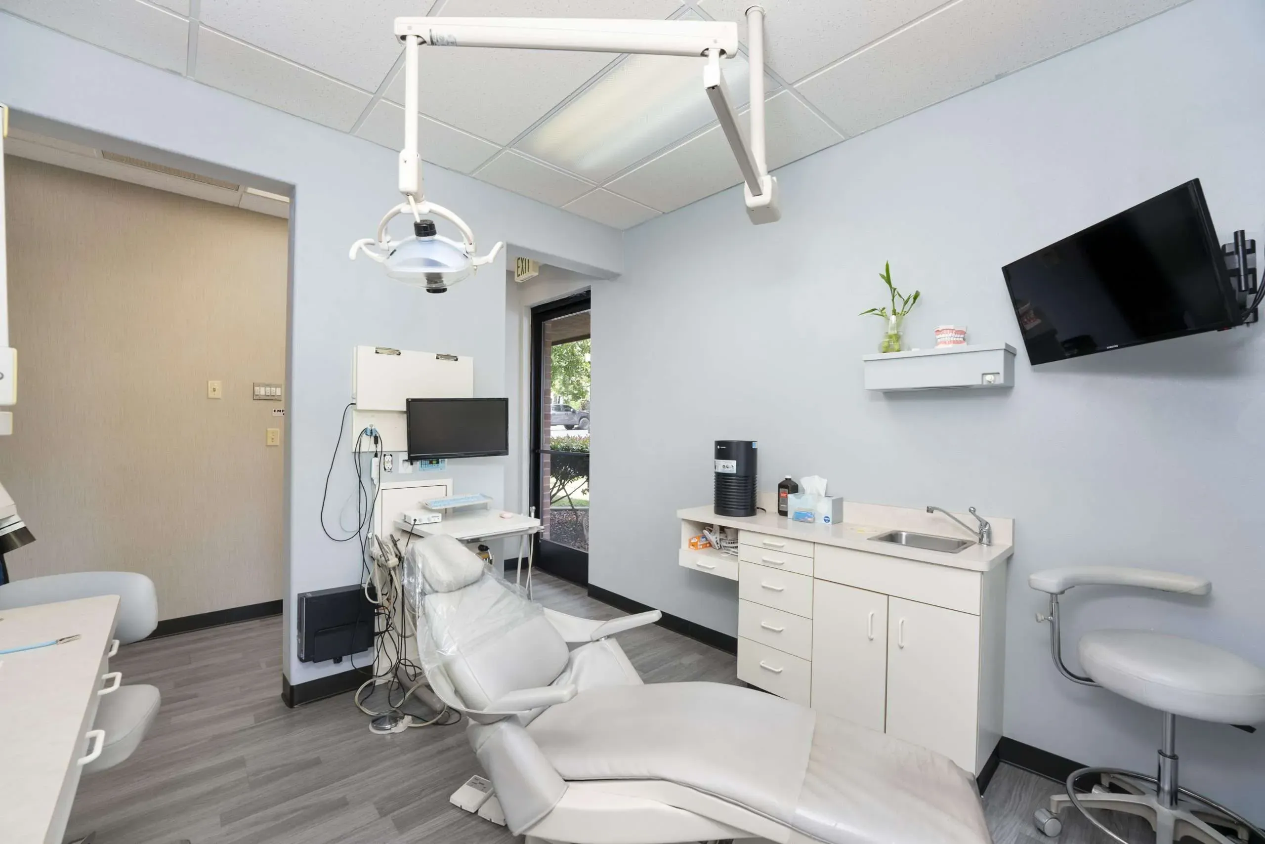 Treatment room with a white dental chair, overhead light, wall monitor and a sink cabinet