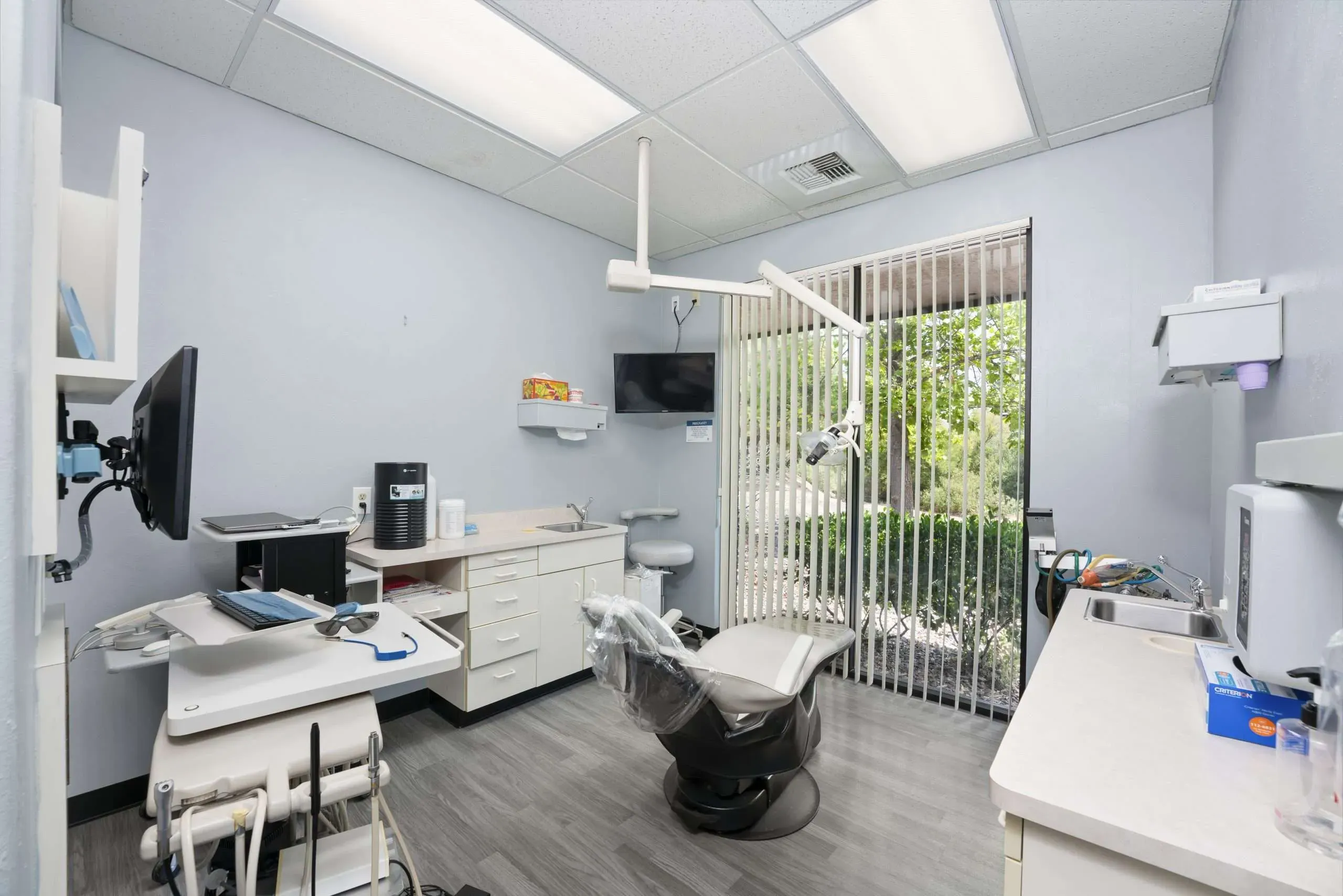 Treatment room with a reclining dental chair, overhead exam light, wall-mounted monitor and a window with blinds